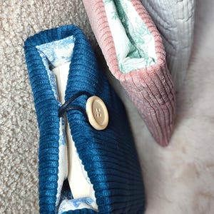May include: Close-up of three corduroy book covers. The front cover is a deep teal blue with a wooden button closure. The interior lining features a blue and white floral pattern. A pink and a gray cover are also visible.