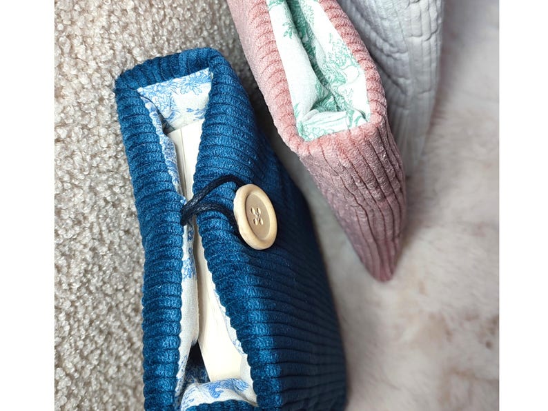 May include: Close-up of three corduroy book covers. The front cover is a deep teal blue with a wooden button closure. The interior lining features a blue and white floral pattern. A pink and a gray cover are also visible.