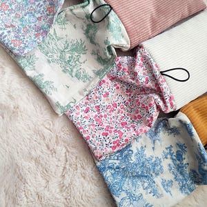 May include: An assortment of fabric pouches in various colors and patterns. The pouches include floral prints in pink and blue, a toile design, and solid colors like teal, pink, and gold. Each pouch has a black loop attached.
