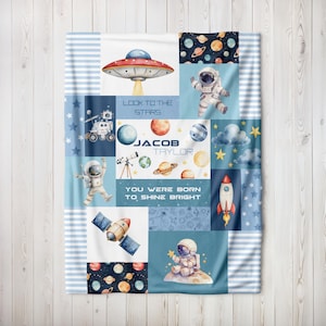 May include: A baby blanket with a space theme, featuring illustrations of planets, astronauts, rockets, and a UFO. The blanket is a patchwork design with the text "Look to the Stars" and "You Were Born to Shine Bright". The name "Jacob Taylor" is also displayed.