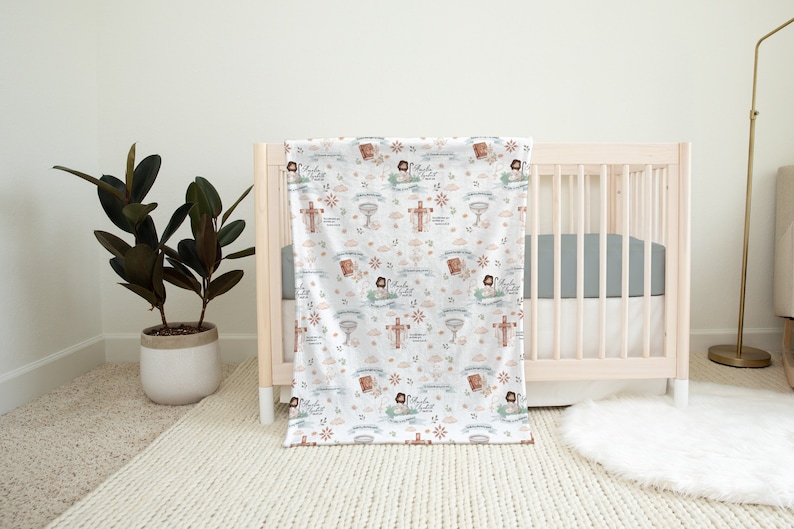 Pu&ograve; includere: Una copertina per neonati bianca con un motivo ripetitivo di immagini religiose, tra cui croci, calici e una figura di Ges&ugrave;. La copertina &egrave; drappeggiata su una culla bianca con un materasso grigio.