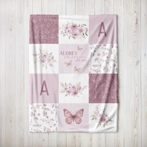May include: A personalized baby blanket with a patchwork design. The blanket features squares with floral arrangements, butterflies, stripes, and the name "Audrey Emerson" with the year "EST. 2023". The color palette includes shades of pink and white.