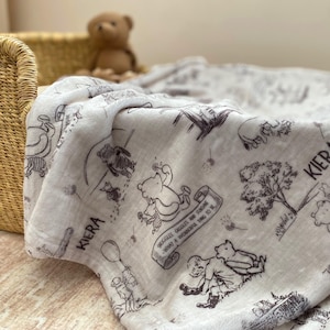 May include: A white muslin baby blanket with a Winnie the Pooh print in black and gray. The blanket features a repeating pattern of Winnie the Pooh and his friends, including Piglet, Tigger, and Eeyore. The blanket is personalized with the name "Kiera" in black lettering. The blanket also features a quote from Winnie the Pooh: "Huggles, giggles, and sunshine, what a wonderful thing to be."