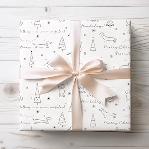 May include: White gift wrap with a black and white dachshund and Christmas tree pattern. The wrapping paper has the text "Happy Howlidays!" and "Merry Christmas Biscuit" repeated on it. The gift is tied with a beige ribbon.