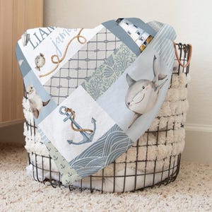 May include: A blue and white baby blanket with a patchwork design featuring a shark, anchor, and seashells. The blanket is folded and placed in a wire basket.