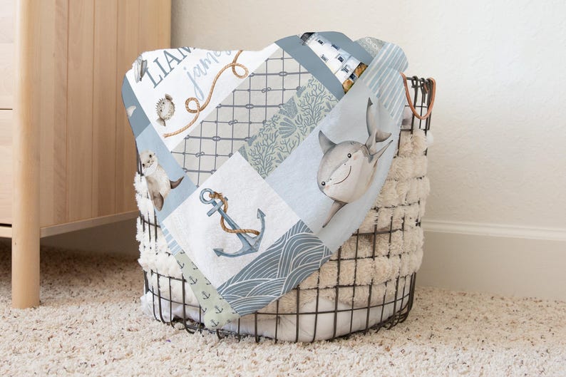 May include: A blue and white baby blanket with a patchwork design featuring a shark, anchor, and seashells. The blanket is folded and placed in a wire basket.