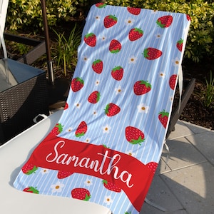 May include: A blue and white striped beach towel with a red strawberry pattern and white flowers. The towel has a red ribbon with the name "Samantha" written in white.