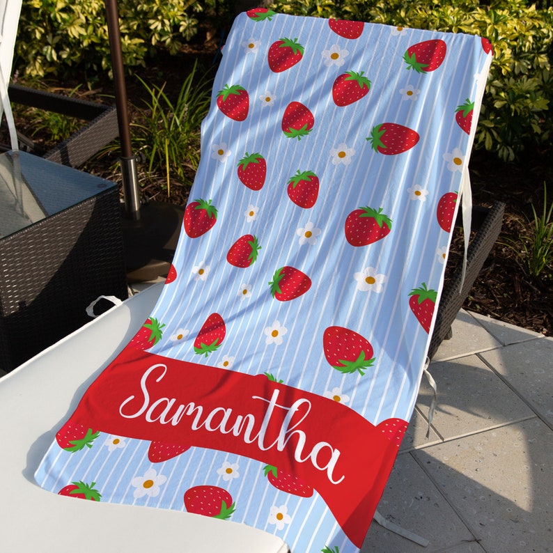 May include: A blue and white striped beach towel with a red strawberry pattern and white flowers. The towel has a red ribbon with the name "Samantha" written in white.