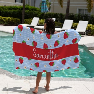 May include: A blue and white beach towel with a red stripe and a strawberry pattern. The towel has the name "Samantha" printed on the red stripe.