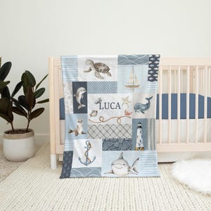 May include: A blue and white baby blanket with a patchwork design featuring sea creatures, a lighthouse, and the name Luca. The blanket is on a white crib with a blue and white striped mattress.