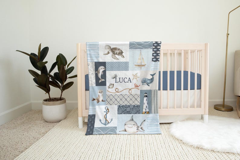 May include: A blue and white baby blanket with a patchwork design featuring sea creatures, a lighthouse, and the name Luca. The blanket is on a white crib with a blue and white striped mattress.