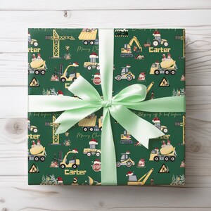 May include: Green wrapping paper with a repeating pattern of construction vehicles, including a crane, dumper truck, cement mixer, and bulldozer. The paper also features a Christmas tree, a stop sign, and the words "Merry Christmas" and "Carter".