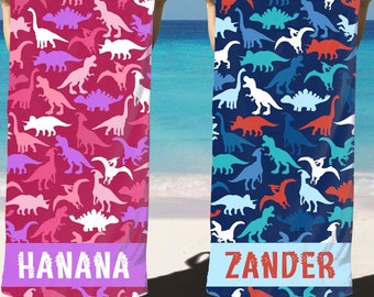 May include: Two colorful beach towels with a dinosaur pattern. The towel on the left is pink with white and purple dinosaurs. The towel on the right is blue with white, red, and blue dinosaurs. The towels have the names "HANANA" and "ZANDER" printed on them.