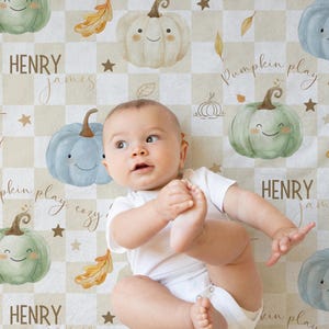 May include: A baby boy lies on a cream and brown checkered play mat with a pumpkin theme. The mat features green and blue pumpkins with smiling faces, fall leaves, and stars. The text "Pumpkin play, cozy days" is written on the mat.