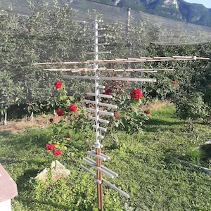 Peut inclure: Un treillis blanc et cuivré dans un jardin, avec des roses rouges et du feuillage vert. Des montagnes sont visibles en arrière-plan sous un ciel bleu. Le treillis est conçu pour soutenir des plantes grimpantes. La structure est en métal.