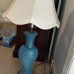May include: A blue ceramic table lamp with a white lampshade. The lampshade has a scalloped edge and a white trim. The lamp base is a vase shape with a gold base.