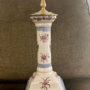 May include: A decorative table lamp with a white ceramic base featuring floral and blue patterns. The lamp has a brass-colored top and a dark brown wooden base. The lamp's design is reminiscent of antique or vintage styles.