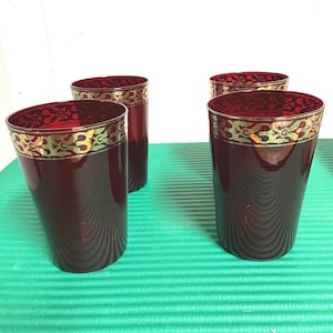 Könnte beinhalten: Vier bordeauxfarbene Trinkgläser mit einem gold-roten Zierrand. Die zylindrischen Gläser scheinen aus Glas zu sein. Die Gläser stehen vor einem grünen Hintergrund.