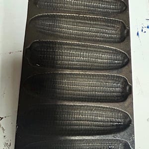 May include: Cast iron cornbread pan with six individual molds shaped like corn cobs. The pan is dark gray and has a handle on one end.