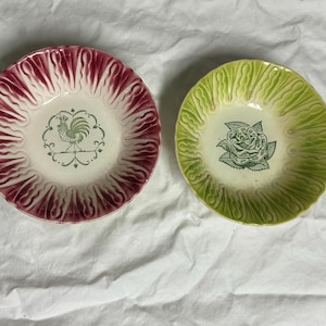 Puede incluir: Dos cuencos decorativos de cerámica. Un cuenco es burdeos con un centro blanco y un diseño de gallo verde. El otro cuenco es verde claro con un centro blanco y un diseño de rosa verde. Ambos cuencos tienen un borde festoneado.