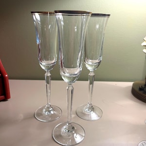 Può includere: Tre flûte da champagne trasparenti con bordi dorati. I bicchieri hanno steli lunghi ed eleganti e basi rotonde. Le flûte sono disposte vicine, mostrando il loro design delicato e il modo in cui catturano la luce.