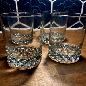 May include: Four clear, short drinking glasses with a textured, bubble-like pattern on the base. The glasses are on a wooden surface, with a dark blue tile background.