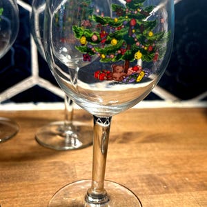 Puede incluir: Copas de vino transparentes con un diseño de árbol de Navidad. El árbol está adornado con adornos, una estrella y regalos. Las copas tienen un tallo y una base transparentes, sobre una superficie de madera.