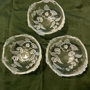May include: Three clear glass bowls with a floral design. Each bowl has a scalloped edge and etched flowers and leaves. The bowls are arranged on a dark green surface.