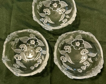 May include: Three clear glass bowls with a floral design. Each bowl features a scalloped edge and etched flowers and leaves. The bowls are arranged on a dark green surface.