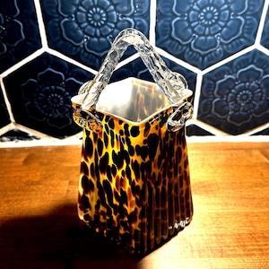 May include: A decorative glass vase with a handle. The vase has a square shape and a pattern of brown and black spots. The handle is clear glass. The vase is sitting on a wooden surface.