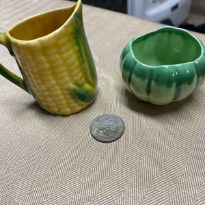 May include: A ceramic corn-shaped creamer and a small green pumpkin-shaped bowl. A silver quarter is next to the bowl. The creamer is yellow and green, and the bowl is a darker green. The items are on a tan herringbone patterned surface.