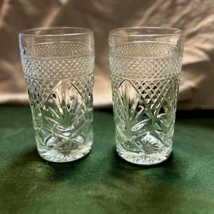 May include: Two clear, patterned drinking glasses. The glasses have a diamond pattern around the top and a leaf-like design in the center. The glasses are sitting on a green surface.
