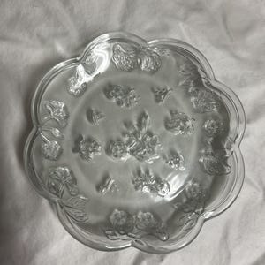 May include: Clear glass serving plate with a scalloped edge and embossed floral design. The plate features detailed rose and leaf patterns, creating a delicate and elegant aesthetic. The plate is round and measures approximately 9 inches in diameter.