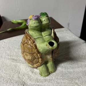 May include: A ceramic teapot shaped like a two-headed turtle. The turtle heads are green with flower accents, and the shell is brown with a textured pattern. The teapot has a spout and a handle, and sits on a white surface.