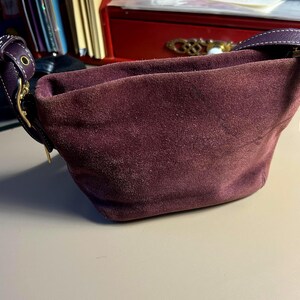 May include: A deep burgundy suede handbag with a curved shape. The bag has a matching burgundy leather strap and gold-toned hardware. The suede has a soft, textured appearance. The bag is designed to be carried over the shoulder.