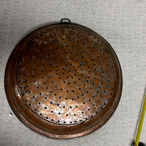 Puede incluir: Un colador redondo de color cobre con numerosos agujeros pequeños. El colador tiene un borde elevado y un pequeño lazo para colgar. El metal muestra signos de edad y uso, con algo de decoloración. Una cinta métrica amarilla está en la esquina.