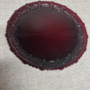 Puede incluir: Un plato redondo de vidrio rojo rubí con un borde decorativo festoneado. El plato presenta un patrón en relieve alrededor del borde. El plato es de un color rojo intenso y rico y parece estar hecho de vidrio.