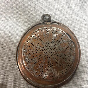 May include: A round, copper-colored kitchen strainer with numerous small holes in a radial pattern. The strainer has a raised rim and a small loop for hanging. The metal has a slightly aged appearance.