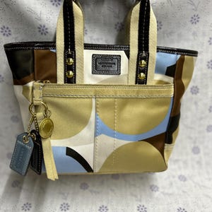 May include: A small, brown, blue, and white patterned Coach tote bag with gold accents and a leather strap. The bag has a Coach logo patch on the front.