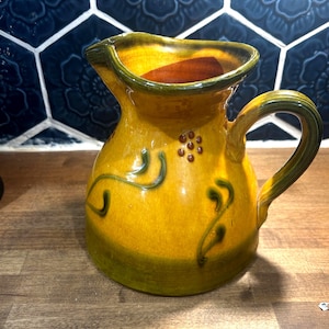May include: A yellow ceramic pitcher with a curved handle and a spout. The pitcher has green accents and a decorative vine design. The interior is a darker shade of yellow. The pitcher is sitting on a wooden surface.