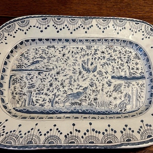 Peut inclure: Un plat rectangulaire en céramique avec un motif peint à la main en bleu et blanc. L'image centrale représente une scène pastorale avec des oiseaux, un cerf et des personnages. La bordure présente un motif répétitif d'arches et de points.