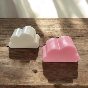 Pode incluir: Dois objetos em forma de nuvem, um em branco sólido e outro em rosa sólido. Cada objeto apresenta uma superfície lisa e arredondada com uma borda superior distinta de três lóbulos, criando uma representação estilizada de uma nuvem.