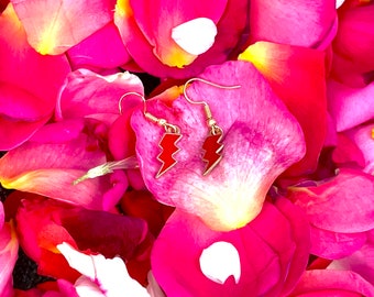 Pode incluir: Um par de brincos de ouro com pingentes de raio vermelho. Os brincos são exibidos em um leito de pétalas de rosa rosa.