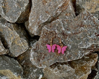 Pode incluir: Um par de brincos de borboleta com glitter rosa e ganchos dourados. Os brincos são fotografados em uma pilha de pedras cinzas.
