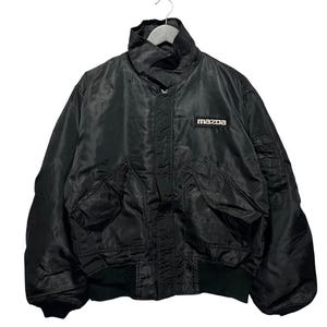Puede incluir: Una chaqueta tipo bomber negra con cuello alto, cierre de cremallera frontal y dos grandes bolsillos con solapa en la parte delantera. La chaqueta tiene un pequeño parche rectangular en el pecho con la palabra Mazda en letras blancas y un bolsillo utilitario.