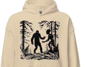 Peut inclure: Un sweat à capuche beige avec un motif noir représentant Bigfoot et un extraterrestre dans une forêt. Le motif est une silhouette, avec les personnages et les arbres en noir sur le fond neutre du sweat.
