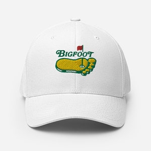 Puede incluir: Gorra de béisbol blanca con una huella de pie bordada en verde y amarillo y el texto "BIGFOOT" en verde. Una bandera roja está en la parte superior de la huella.