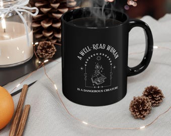 Può includere: Tazza in ceramica nera con la scritta bianca "A WELL-READ WOMAN IS A DANGEROUS CREATURE". La tazza presenta un design a mezzaluna e stelle con un libro aperto ed elementi floreali. Vapore che esce dalla tazza.