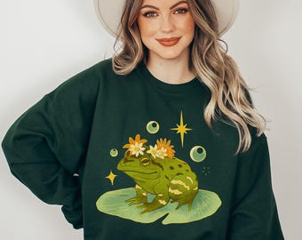 Puede incluir: Una sudadera verde oscuro con un gráfico de una rana con una corona de flores. La rana está sentada en una hoja de nenúfar con burbujas y una estrella alrededor.