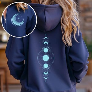 May include: A navy blue zippered hoodie with a moon and stars design on the left side and a moon phase design on the right side. The text "find beauty in every phase" is printed on the left side of the hoodie.